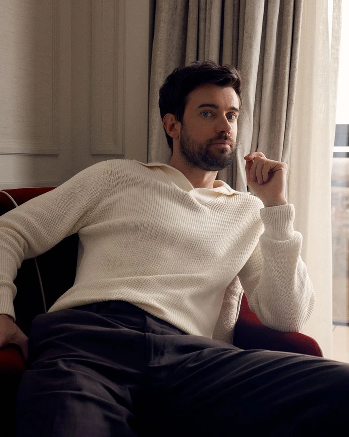 Jack Whitehall seated in a red armchair wearing a cream polo sweater and dark trousers near a draped window.