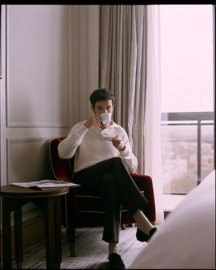 A man seated in a red armchair in a hotel room, drinking from a white teacup while holding a saucer.