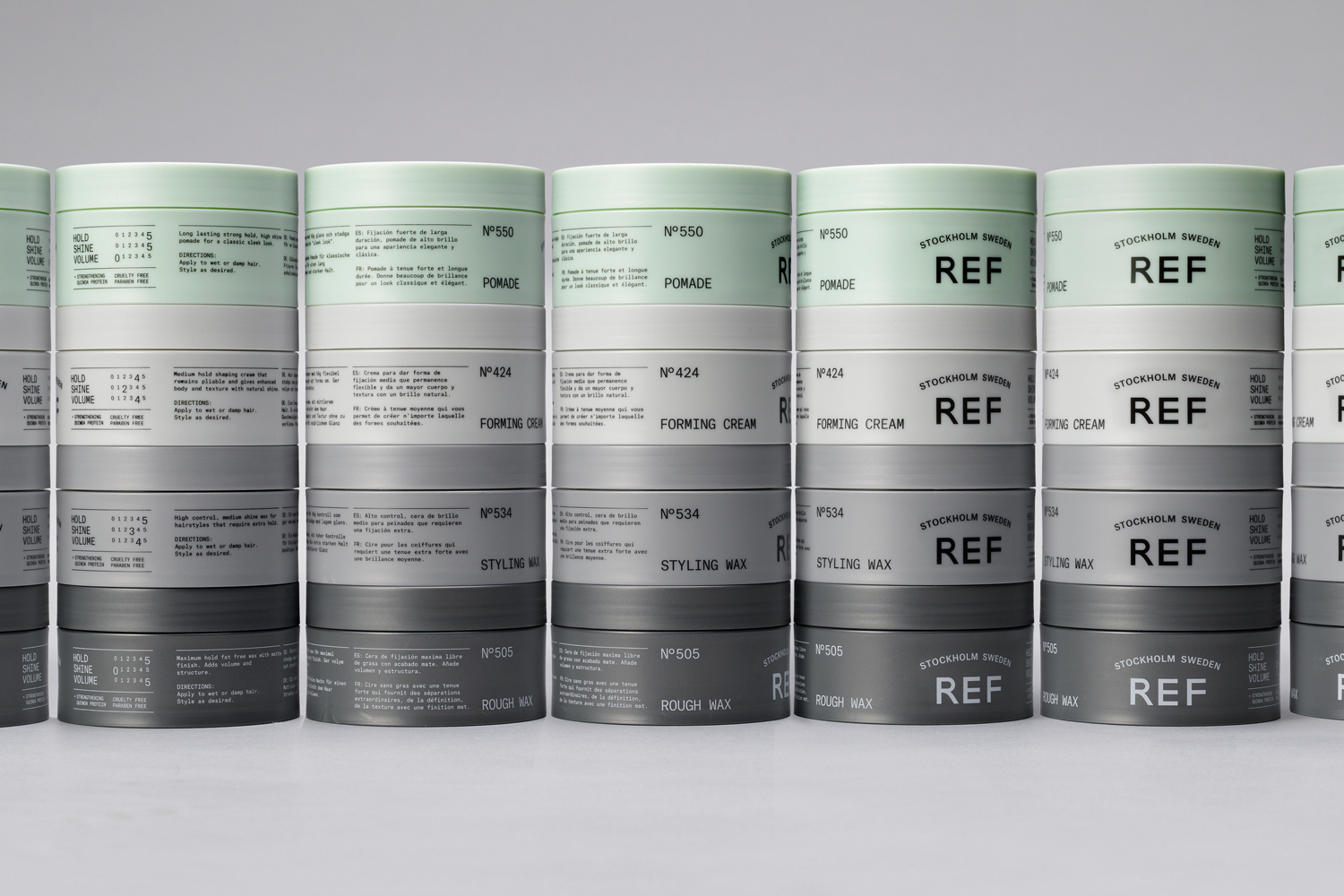 Stacks of REF hair styling product containers in varying heights and muted shades against a grey background.