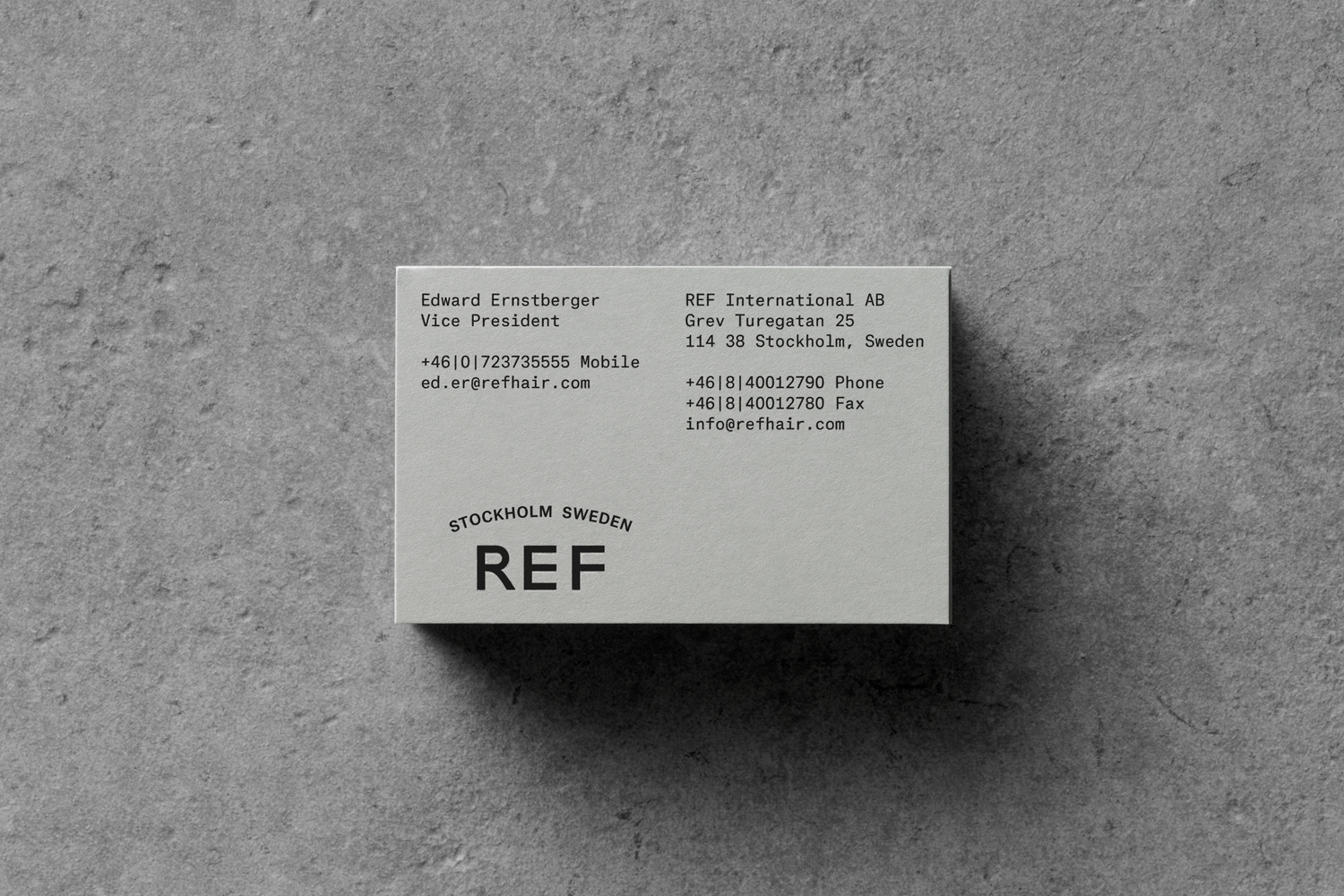 Top-down view of a minimalist white business card for REF resting on a textured grey concrete surface.
