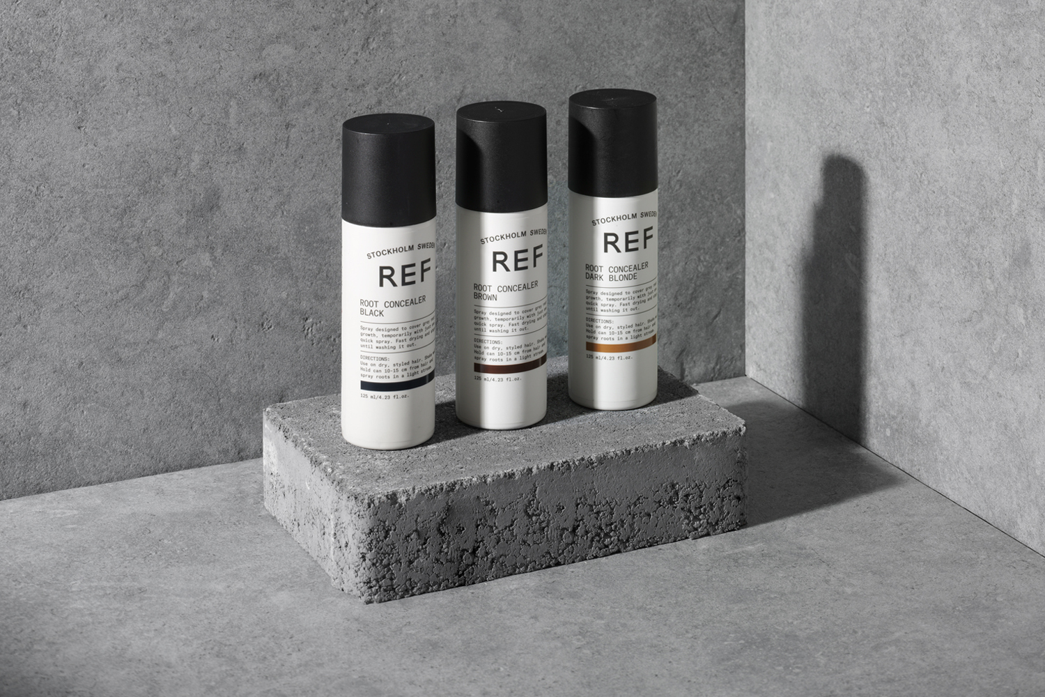 Three REF root concealer spray bottles displayed upright on a textured concrete block against a grey wall