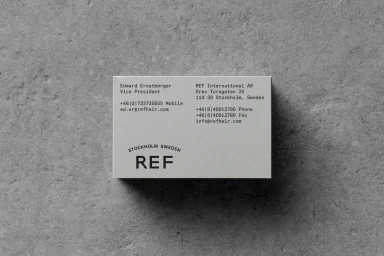 Top-down view of a minimalist white business card for REF resting on a textured grey concrete surface.