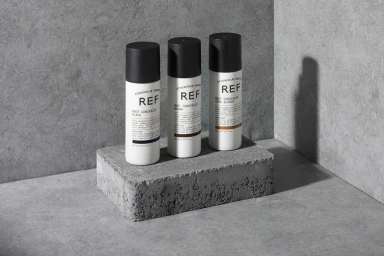 Three REF root concealer spray bottles displayed upright on a textured concrete block against a grey wall