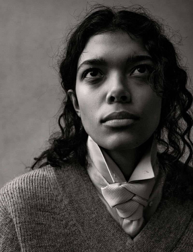Portrait of a model with dark curly hair wearing a textured v-neck sweater, collared shirt, and knotted tie against a neutral background