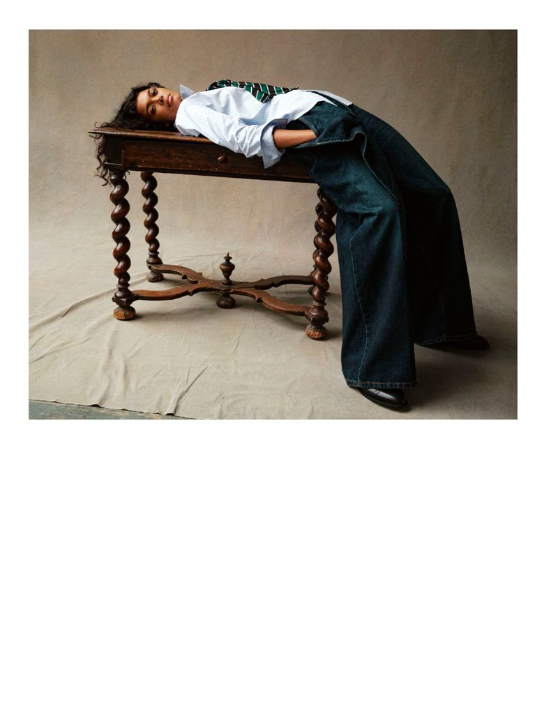 Model lying horizontally across an antique wooden table wearing a white shirt and wide-leg denim pants for Dapper Dan Magazine.
