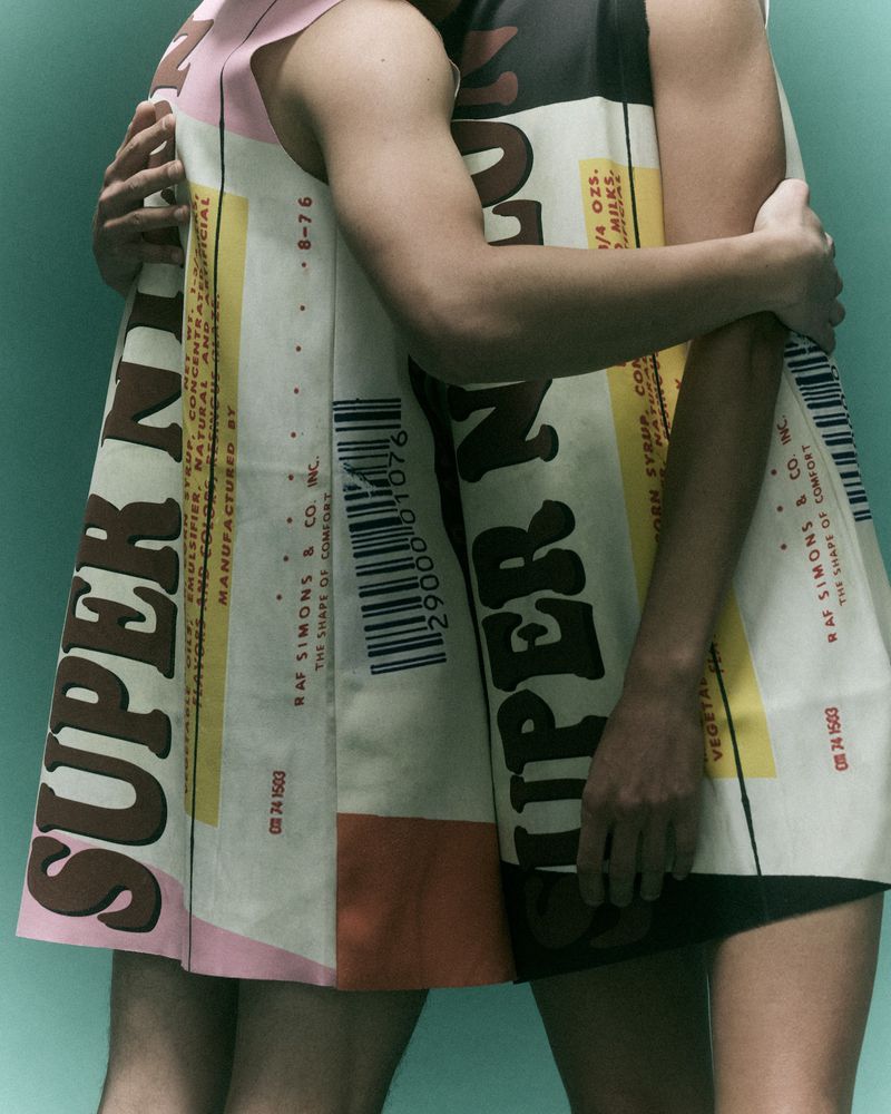Two models wearing printed garments with typographic patterns embracing against a solid teal studio background.