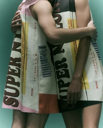 Two models wearing printed garments with typographic patterns embracing against a solid teal studio background.