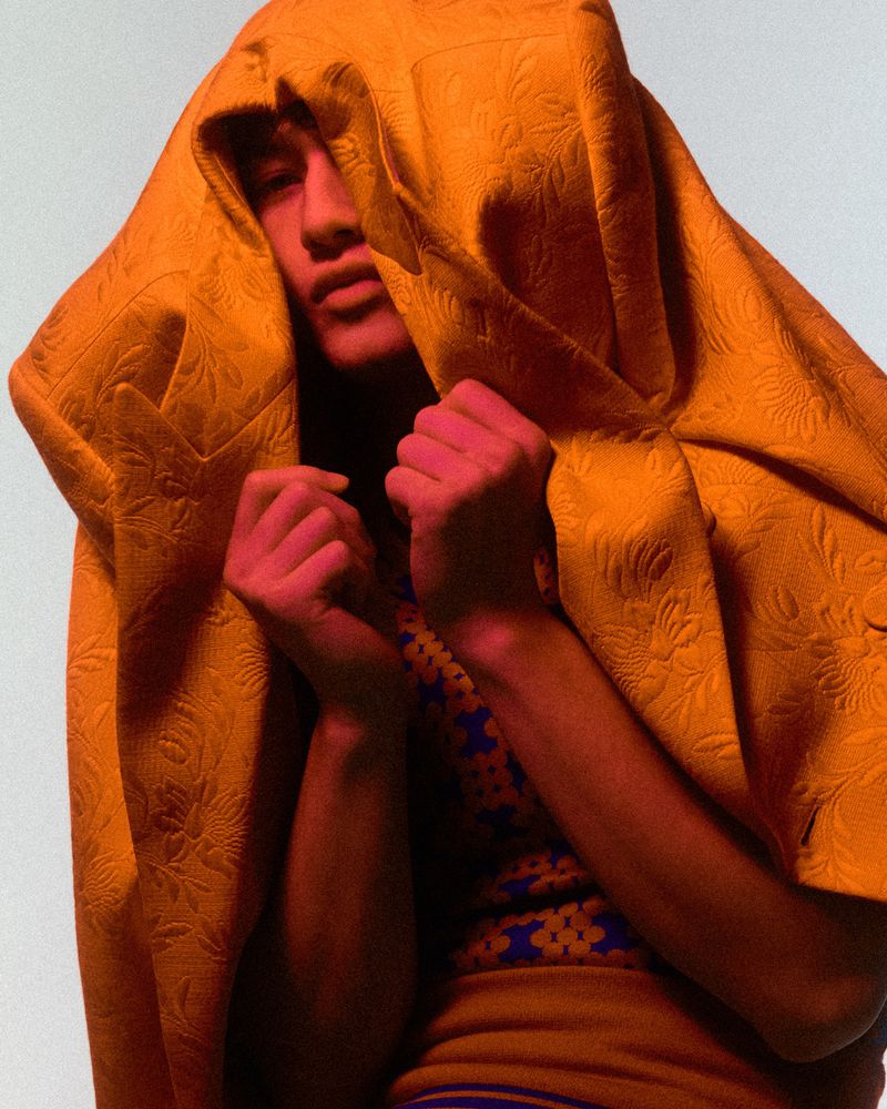 Model with head draped under a yellow patterned fabric holding fists up against a neutral background for Behind the Blinds