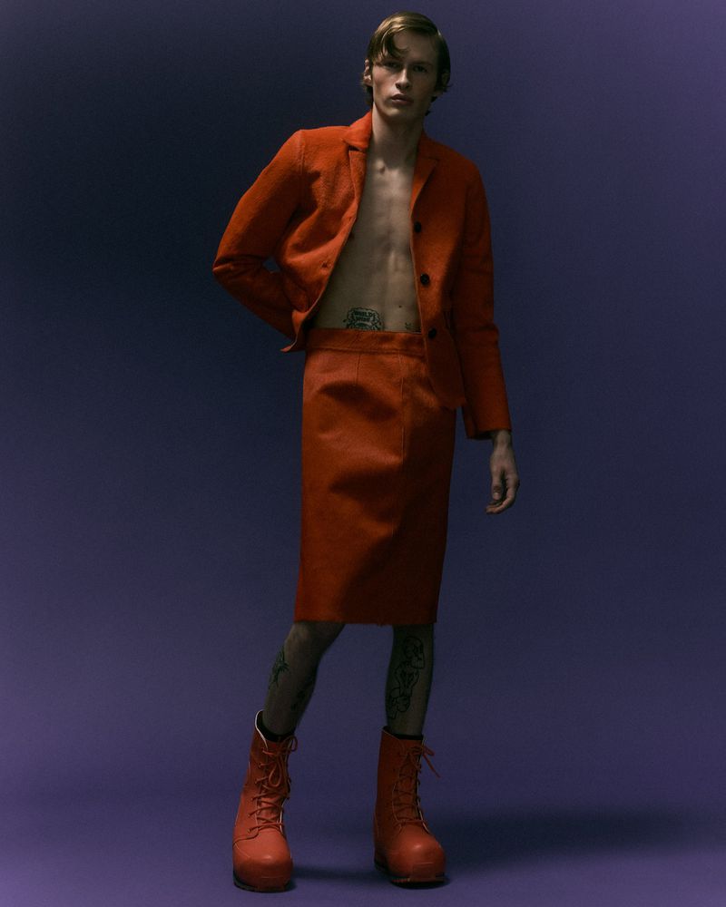 Young man posing in an orange suit jacket, matching skirt, and boots against a dark purple background.
