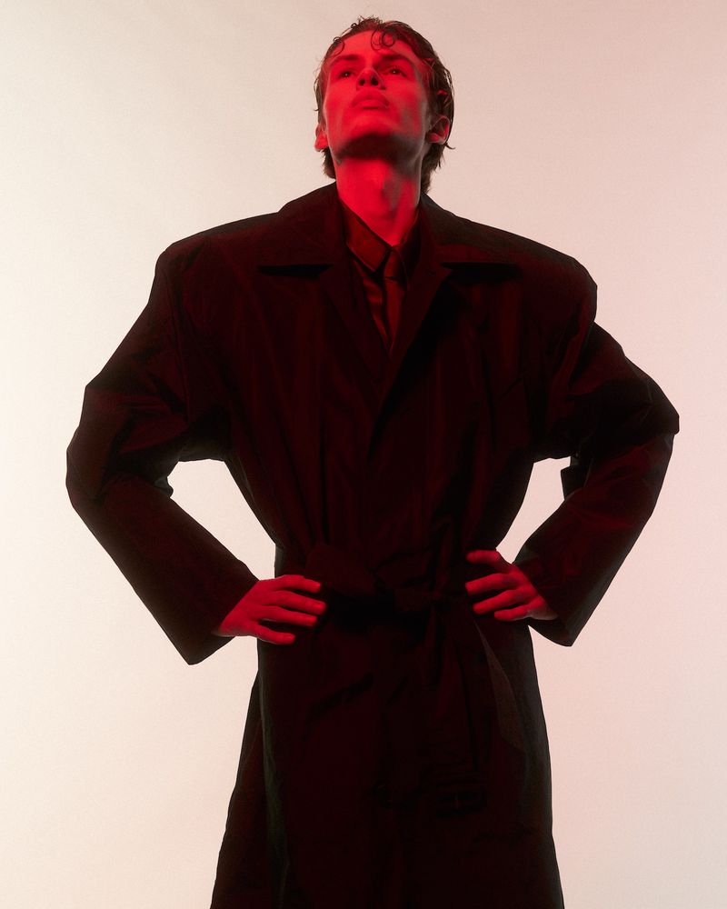 Model wearing an oversized dark coat with hands on hips under red lighting against a light background for Behind the Blinds.