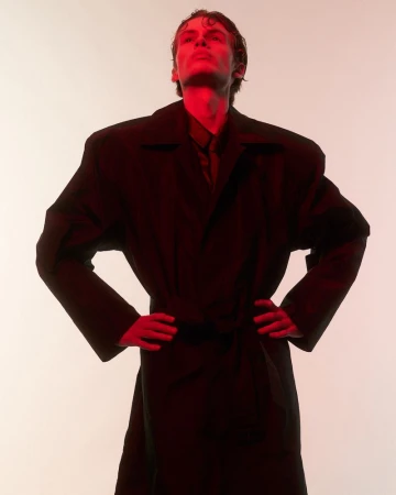 Model wearing an oversized dark coat with hands on hips under red lighting against a light background for Behind the Blinds.