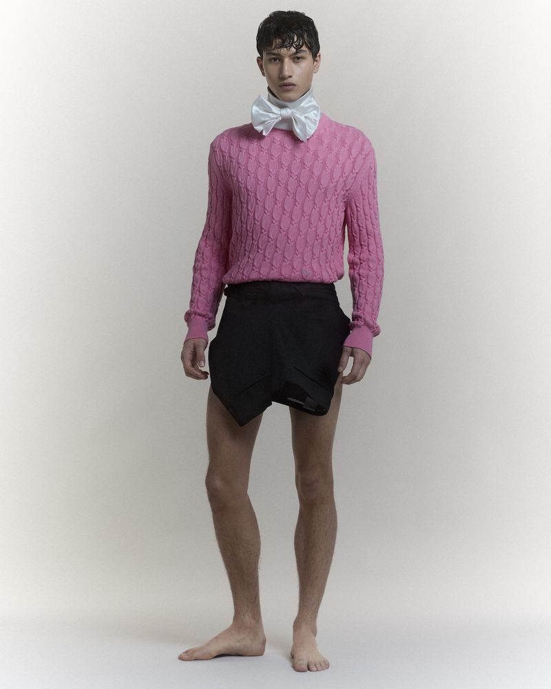 Model wearing a pink cable-knit sweater, white bow tie, and dark shorts against a seamless grey backdrop.