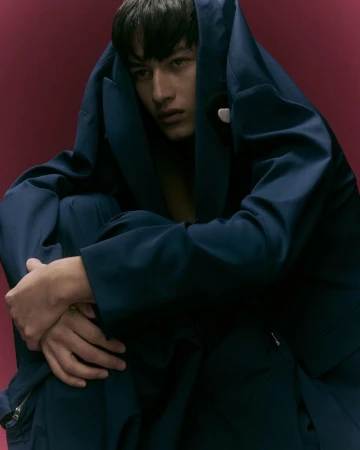Young male model crouching in a blue suit with jacket draped over his head against a dark red background.