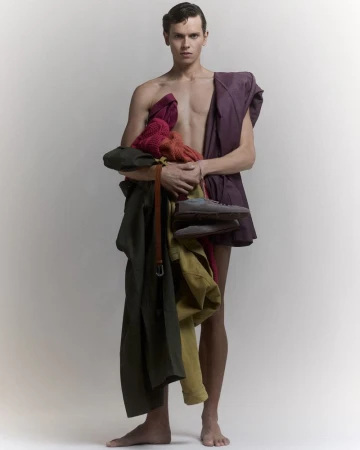 Shirtless male model holding a stack of layered garments and shoes against a plain white studio background.