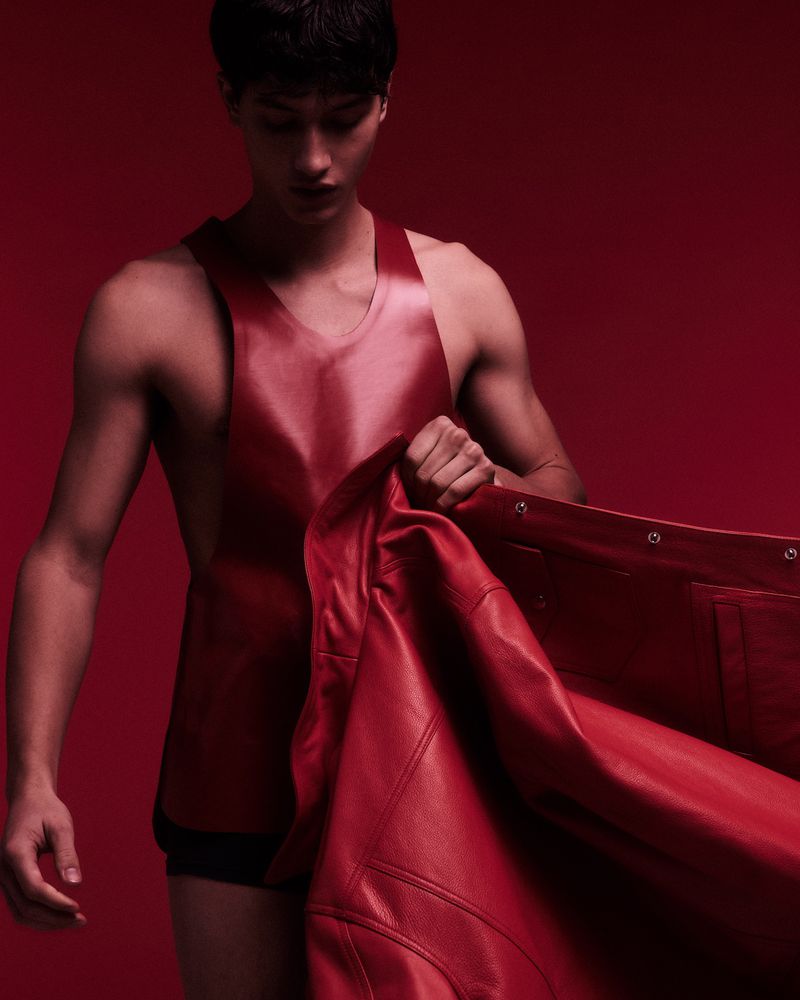 A young man wearing a red leather top holding a matching red jacket against a solid dark red background