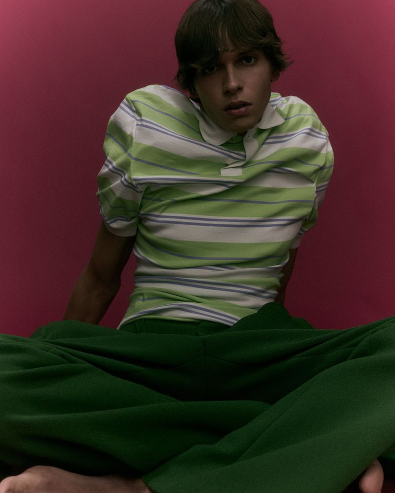 Model seated wearing a green striped polo shirt and dark green trousers against a solid dark pink background.