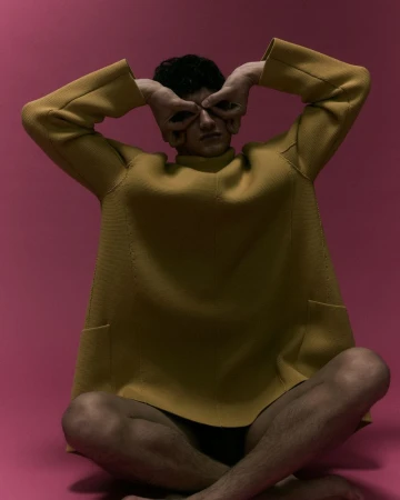 Model seated on the floor wearing a yellow tunic with hands forming binoculars around eyes against a pink backdrop