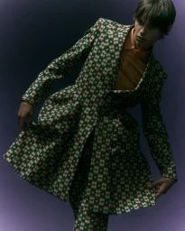 Model wearing a patterned green coat and orange shirt against a dark purple background for Behind the Blinds