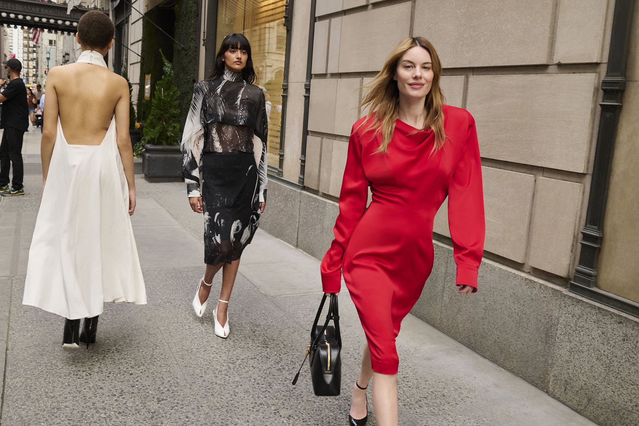 Three models walking on a city sidewalk wearing stylish dresses, photographed by Angelo Pennetta for Saks Fifth Avenue.