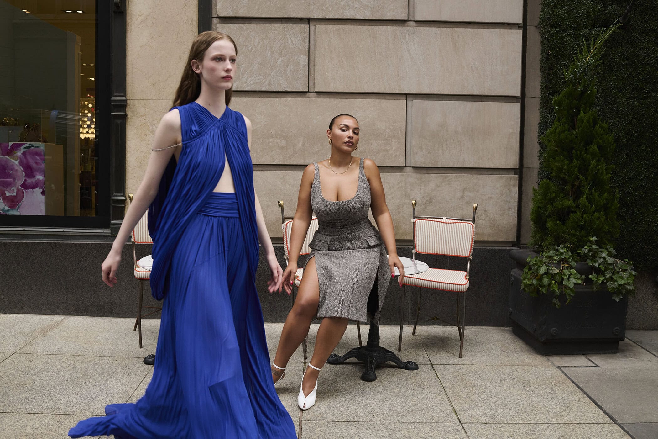 Two female models posing outside, one in a flowing blue pleated gown and one seated wearing a grey skirt suit