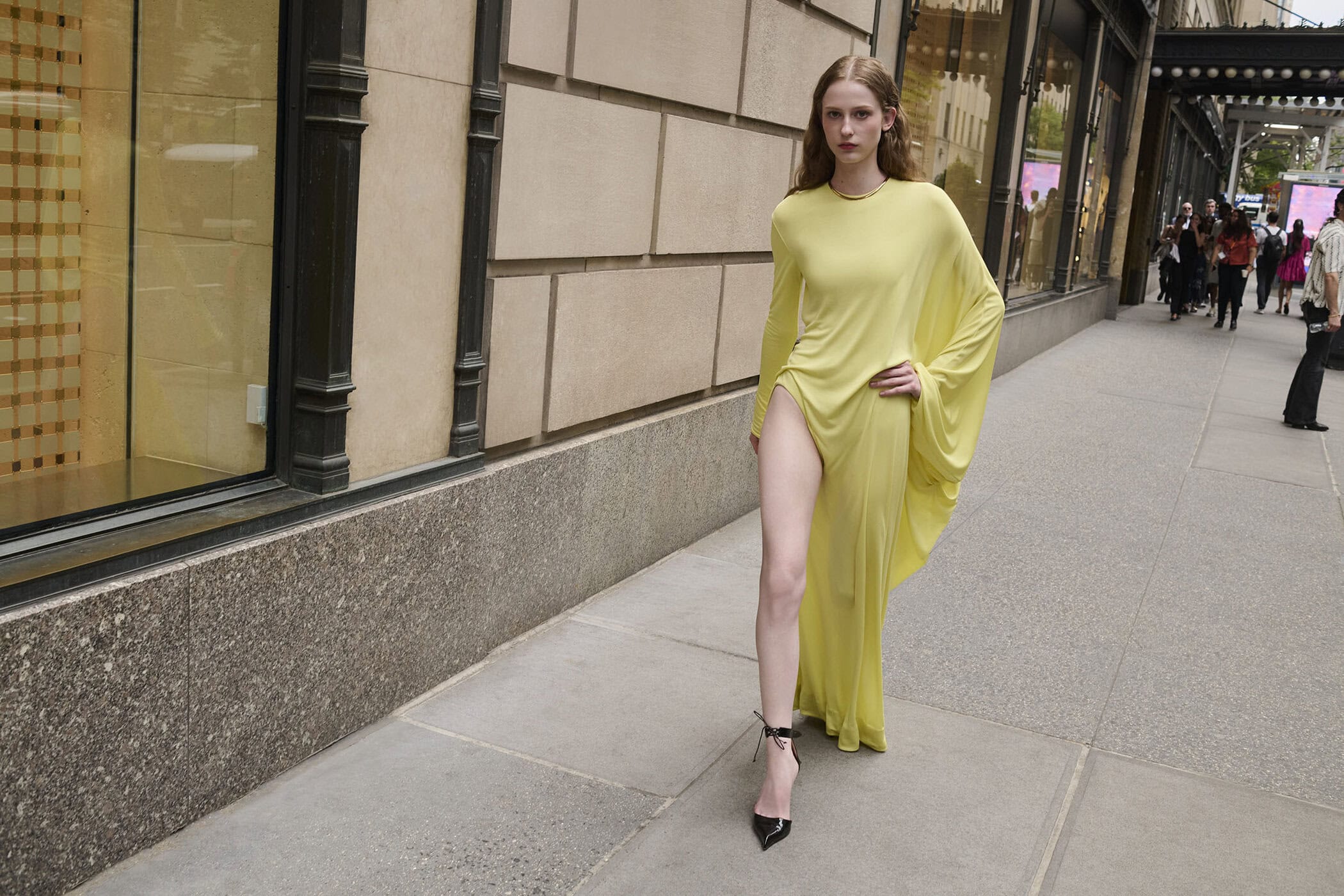 Model in a yellow dress walking on a city sidewalk next to a stone building facade
