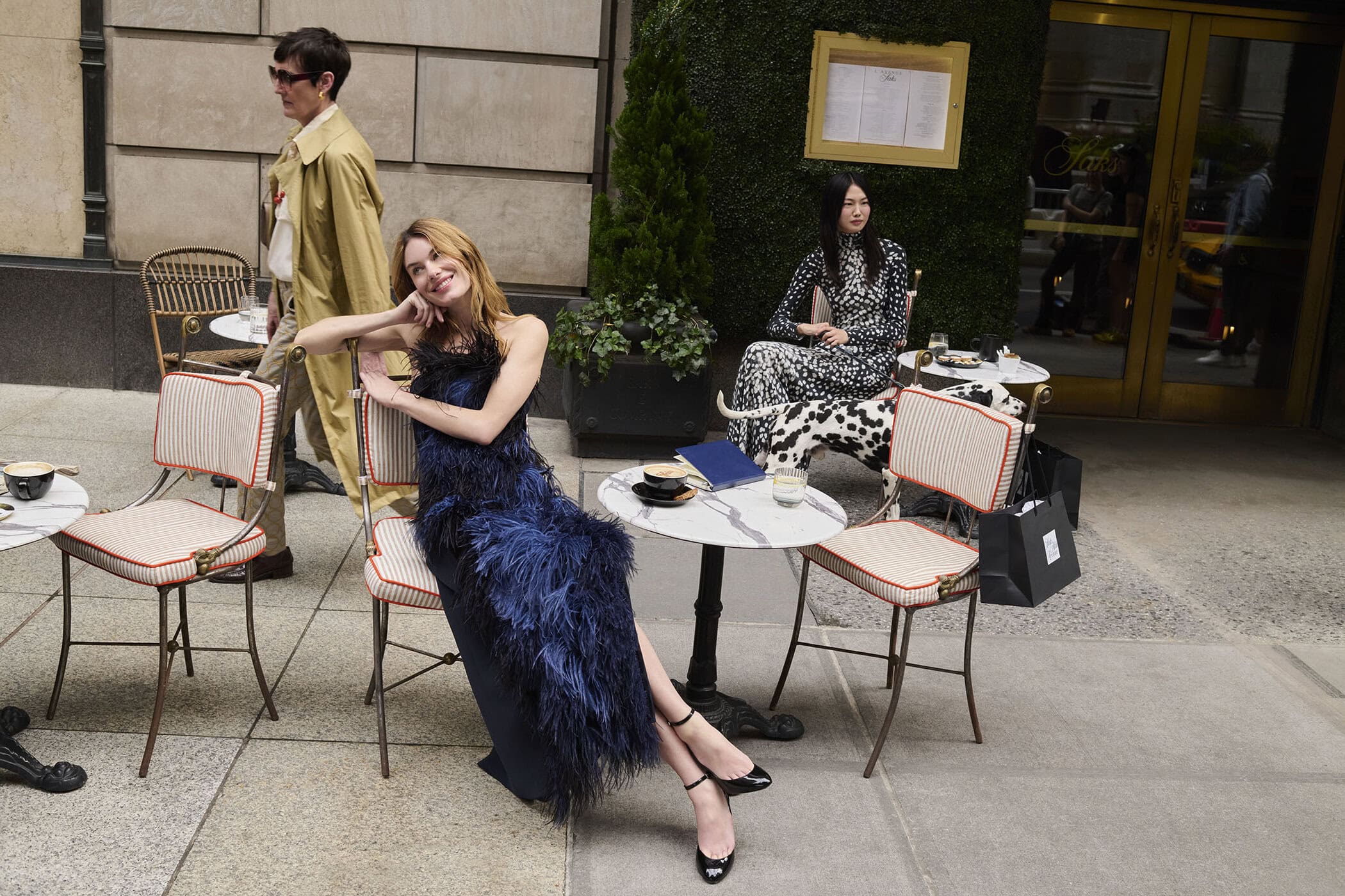 Model seated outside at a cafe table wearing a dark feather-trimmed gown for Saks Fifth Avenue Fall 2025 campaign.