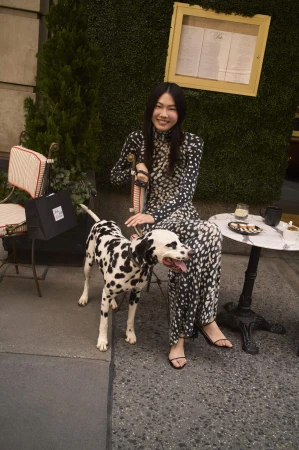 Woman in printed turtleneck dress seated at an outdoor cafe table with a Dalmatian dog for Saks Fifth Avenue Fall 2025 campaign