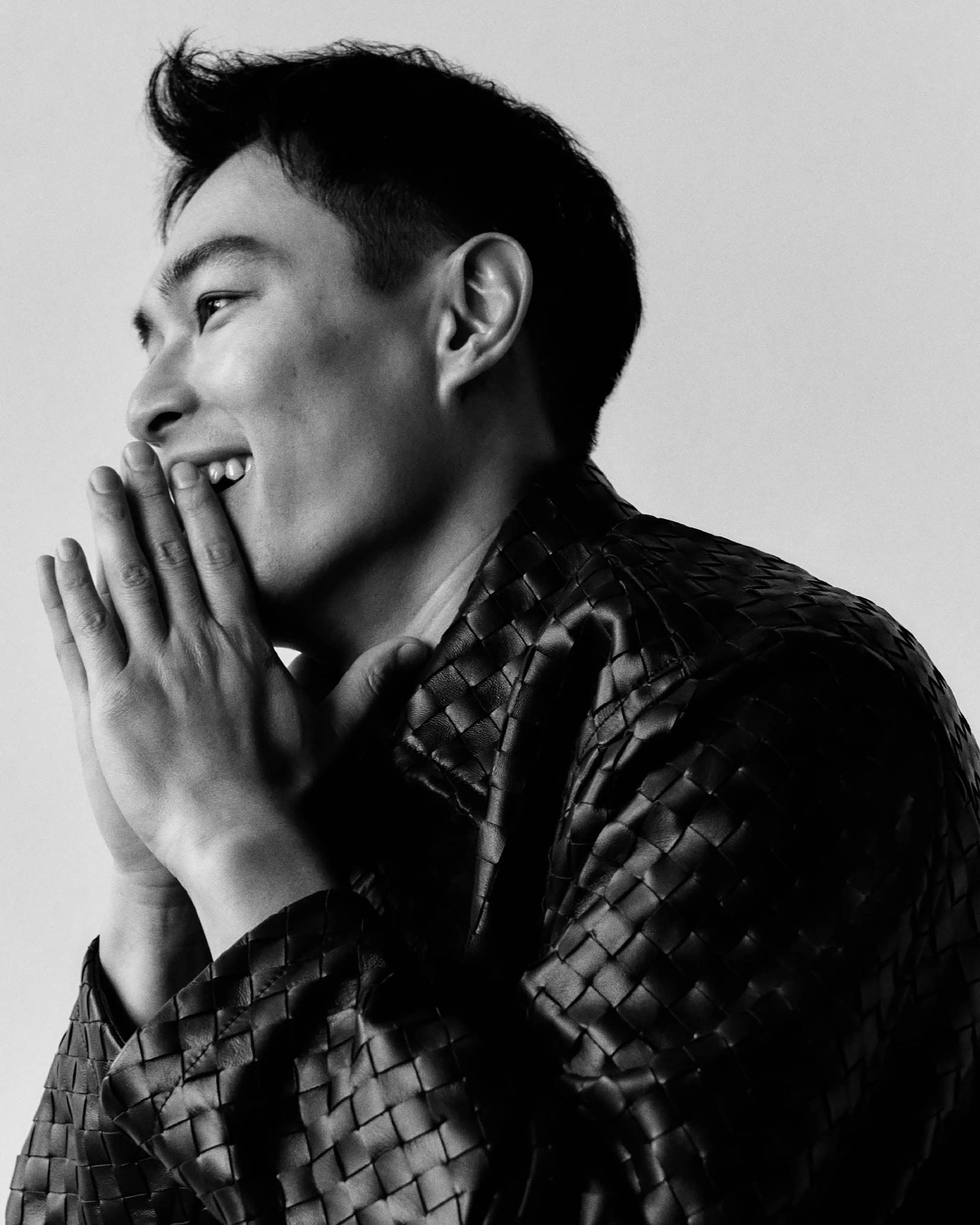 Monochrome portrait of smiling man wearing a woven leather jacket with hands clasped near face against a plain light background