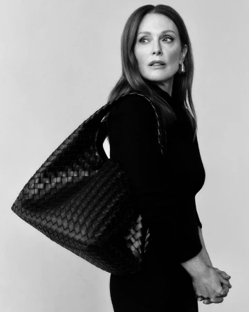 Julianne Moore wearing black clothing holding a woven leather handbag against a neutral background