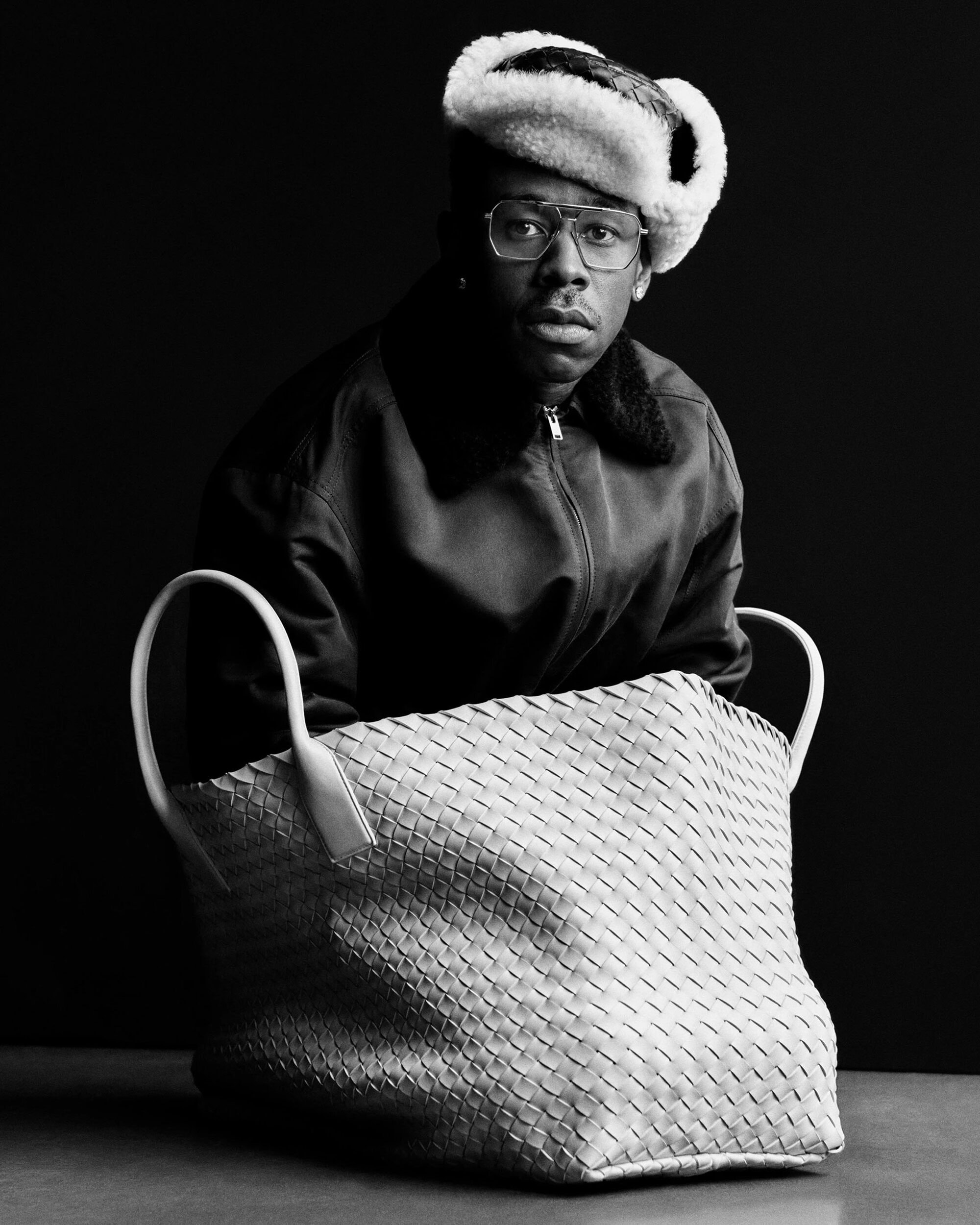 Monochrome portrait of a man wearing glasses and a fur hat behind a large woven leather bag for Bottega Veneta