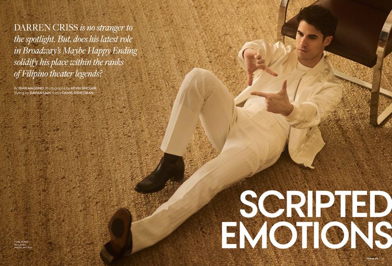 Darren Criss lies on a carpet, wearing a white suit, pointing his fingers to create a rectangle frame. He wears black boots.