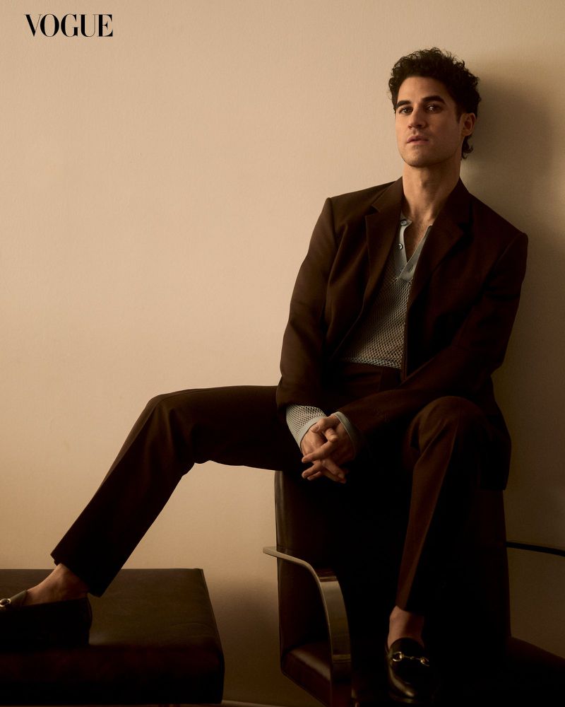 A fashionable man in a brown suit sits in a studio with his hands clasped, with the word 'Vogue' visible in the corner of the frame.