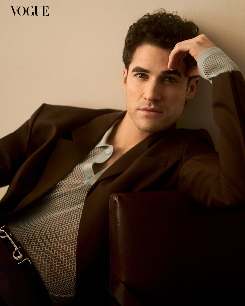 Darren Criss poses in a brown suit jacket and sheer mesh shirt in a studio shot for Vogue. He leans on a brown leather chair.