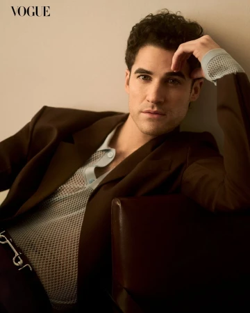Darren Criss poses in a brown suit jacket and sheer mesh shirt in a studio shot for Vogue. He leans on a brown leather chair.