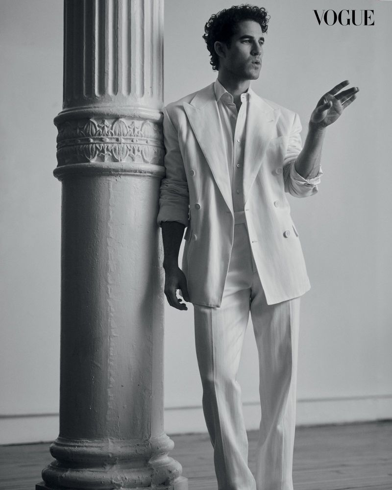 Portrait of Darren Criss in a white suit leaning against a fluted column in a studio, with the Vogue logo visible in the upper right.