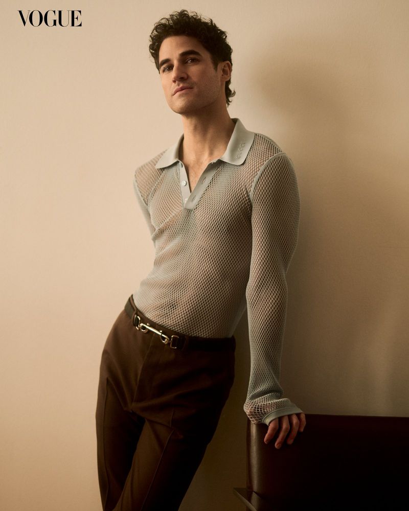 Darren Criss poses indoors wearing a semi-sheer polo shirt and brown trousers, standing next to a dark chair, Vogue logo visible.