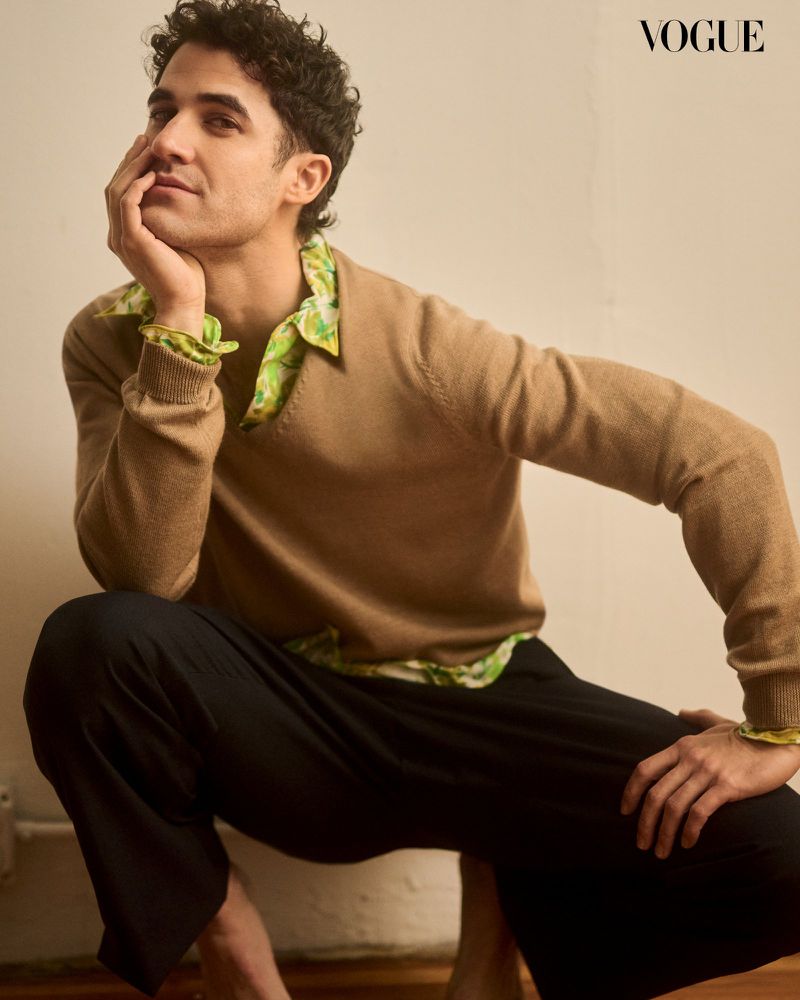 Male model with curly hair posing in a tan sweater and black pants, Vogue logo visible in the top right corner, indoor setting.