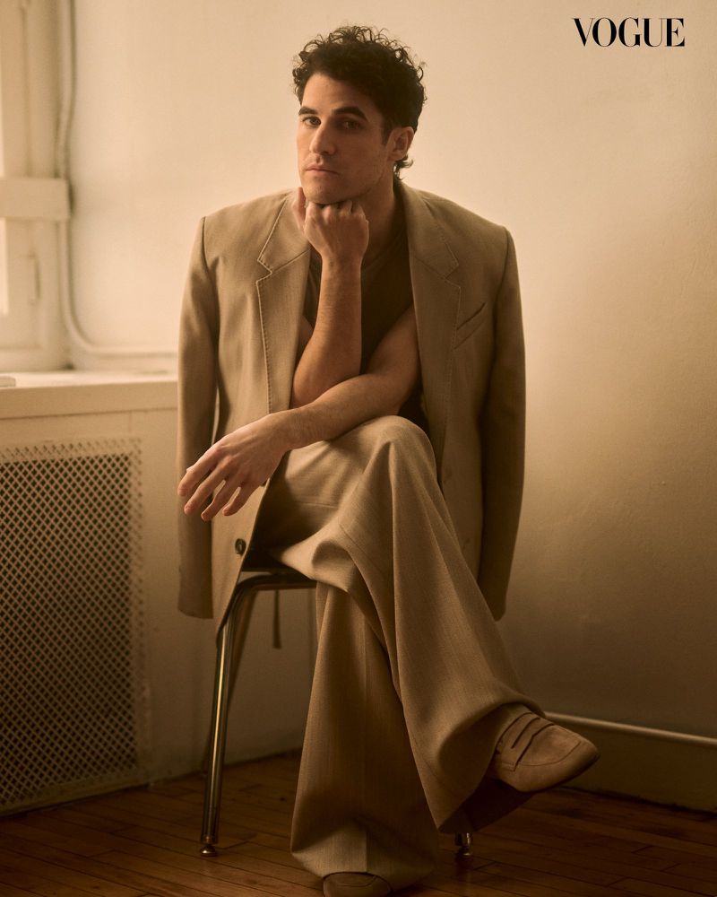 A handsome man sits on a chair in a stylish beige suit, looking thoughtfully at the camera in a minimalist indoor setting.