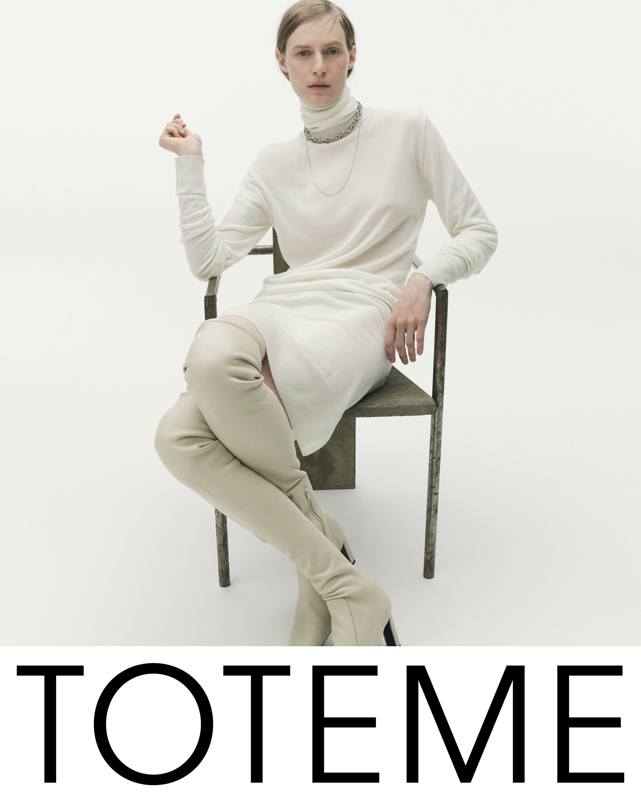 Model Julia Nobis seated in an armchair wearing a cream turtleneck dress and knee-high boots for Toteme Fall 2025 campaign.