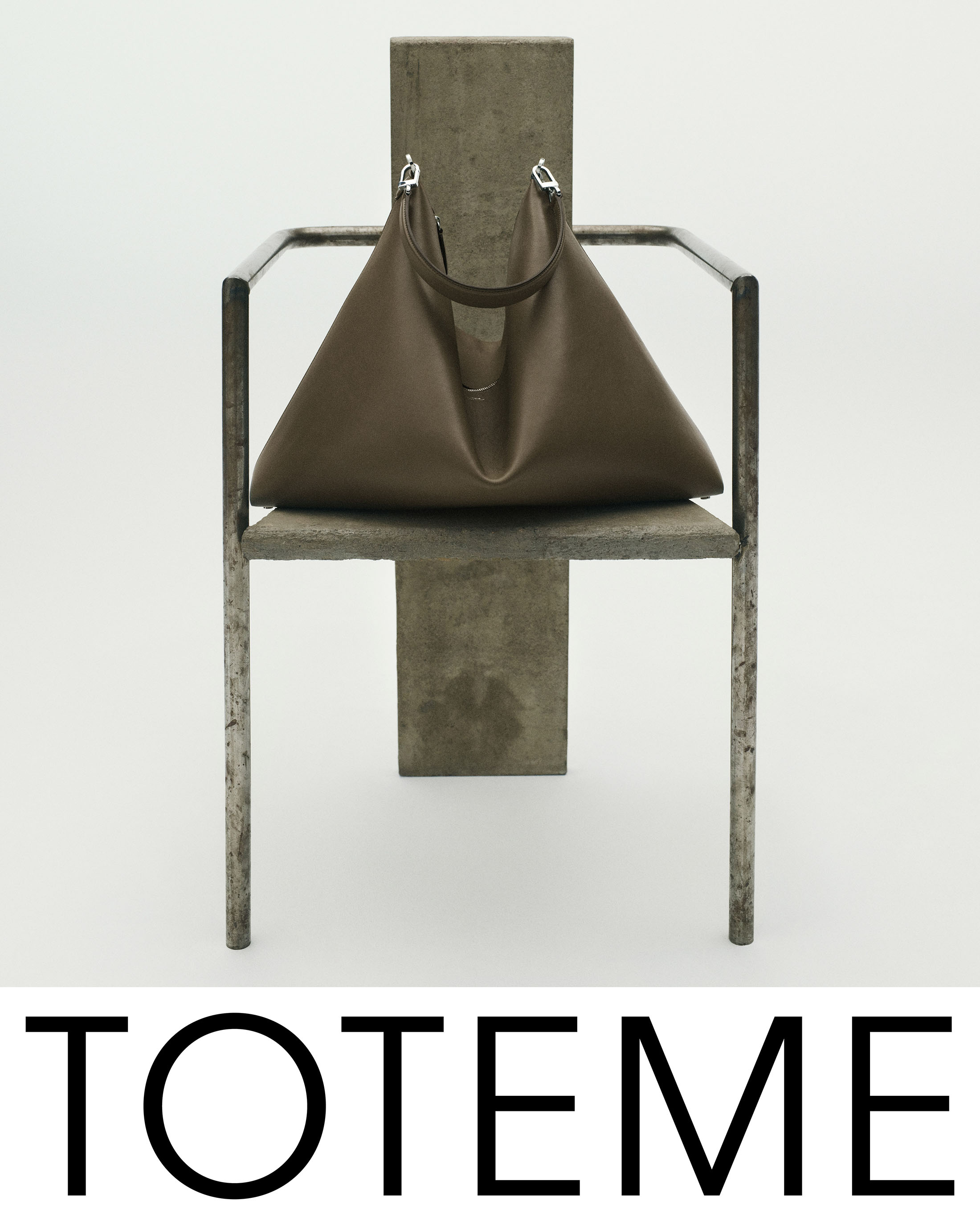 Brown leather handbag resting on a sculptural metal and stone chair against a plain white background for Toteme Fall 2025 ad campaign.