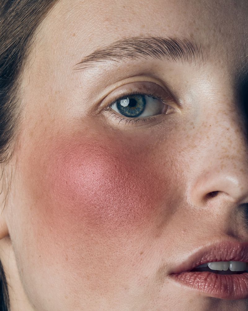 Close-up portrait of a woman's face highlighting detailed skin texture, freckles, and striking flush of pink blush across her cheek.