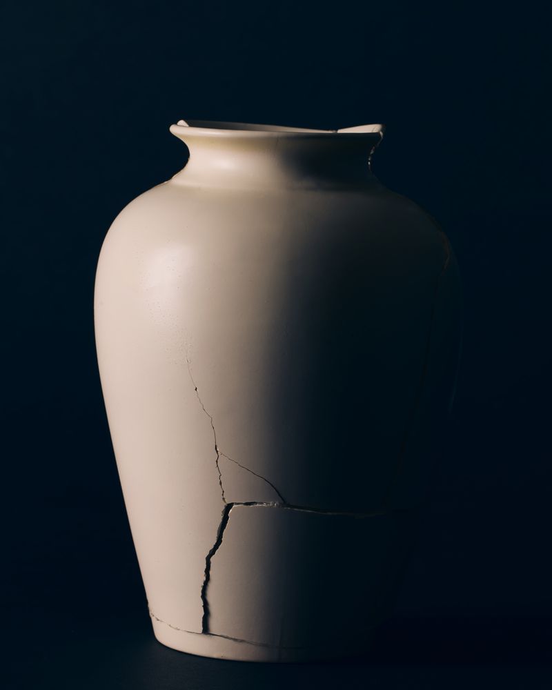 A cracked white ceramic vase stands centrally illuminated against a dark background in low-key studio lighting.