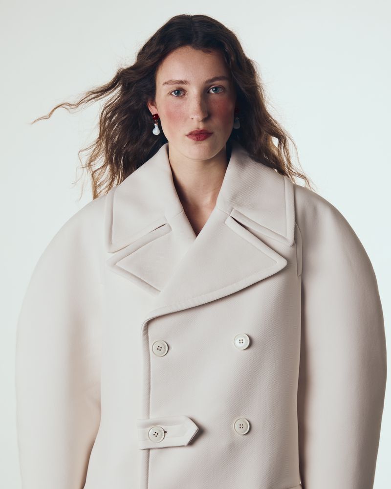 Model with long brown hair wearing a cream double-breasted coat against a plain light background for Vogue Portugal.