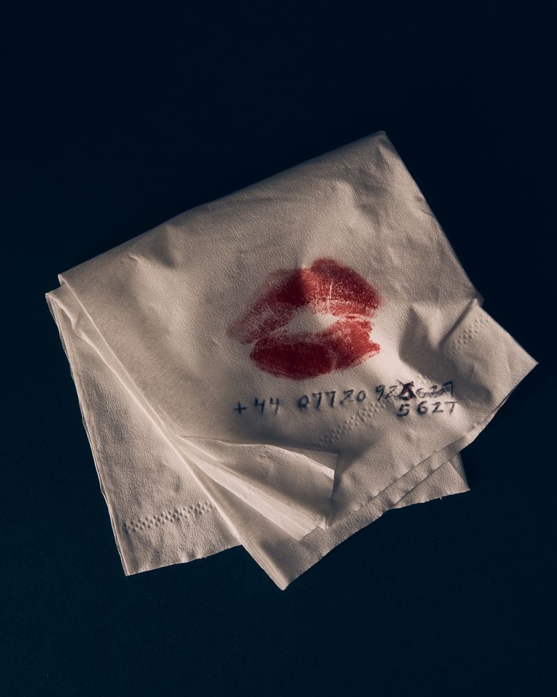 Folded white paper napkin with red lipstick kiss mark and handwritten phone number on dark background