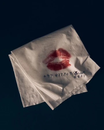 Folded white paper napkin with red lipstick kiss mark and handwritten phone number on dark background