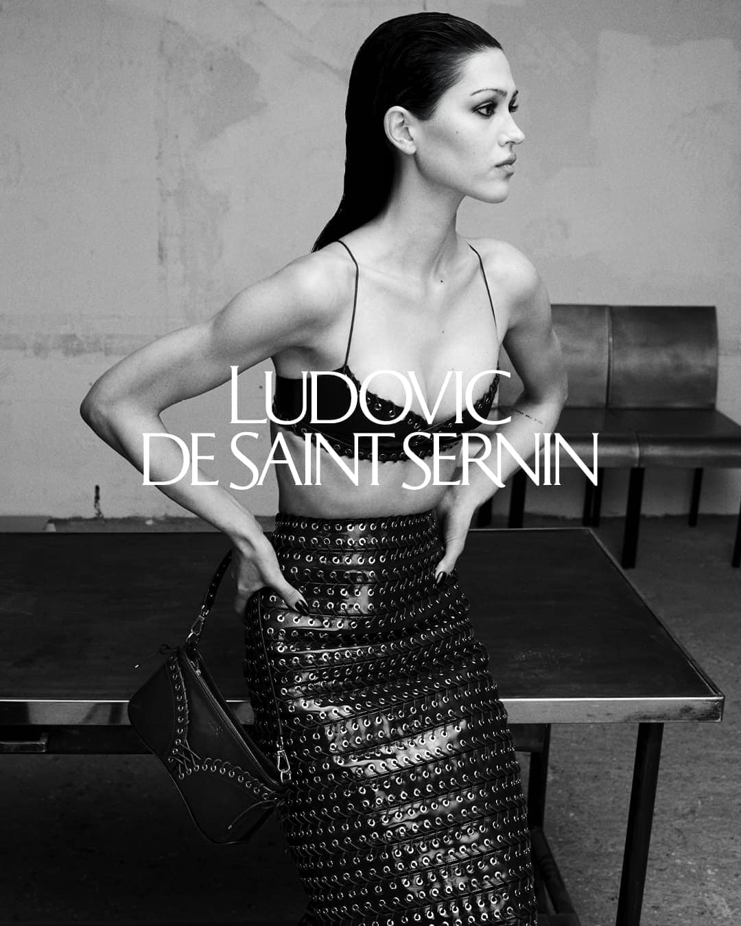 Monochrome fashion campaign image featuring a model wearing a dark laced bralette and patterned skirt against an interior backdrop.