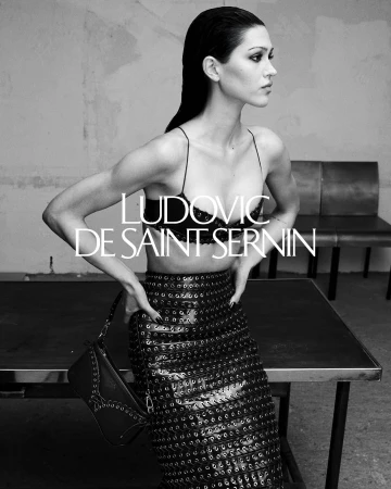 Monochrome fashion campaign image featuring a model wearing a dark laced bralette and patterned skirt against an interior backdrop.