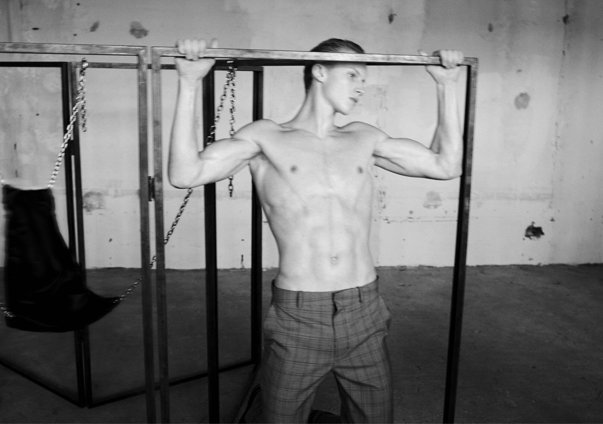 Shirtless man wearing plaid trousers holding onto a metal framing structure in an industrial room