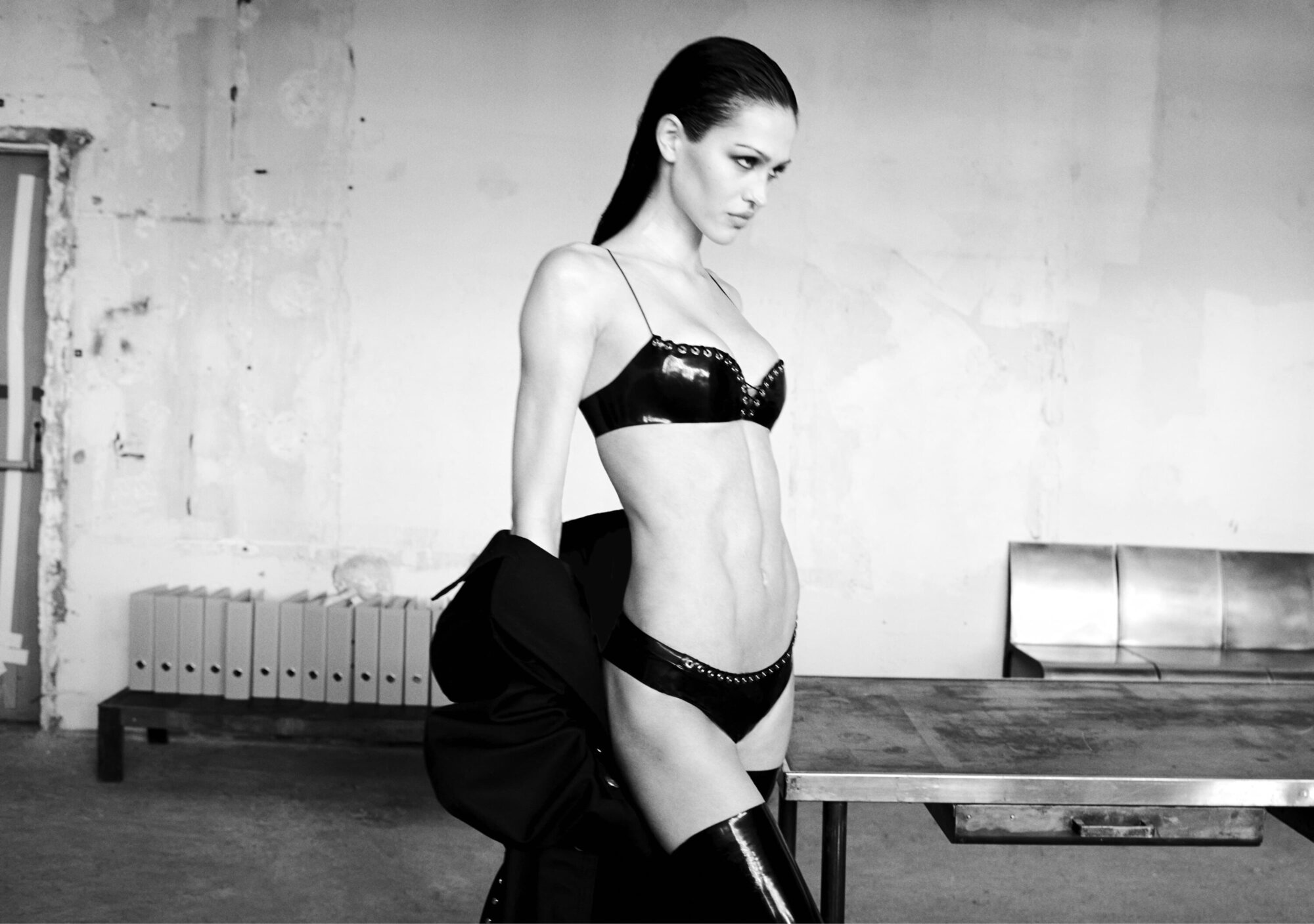 Model wearing dark undergarments and knee-high boots standing in an industrial room with a metal table