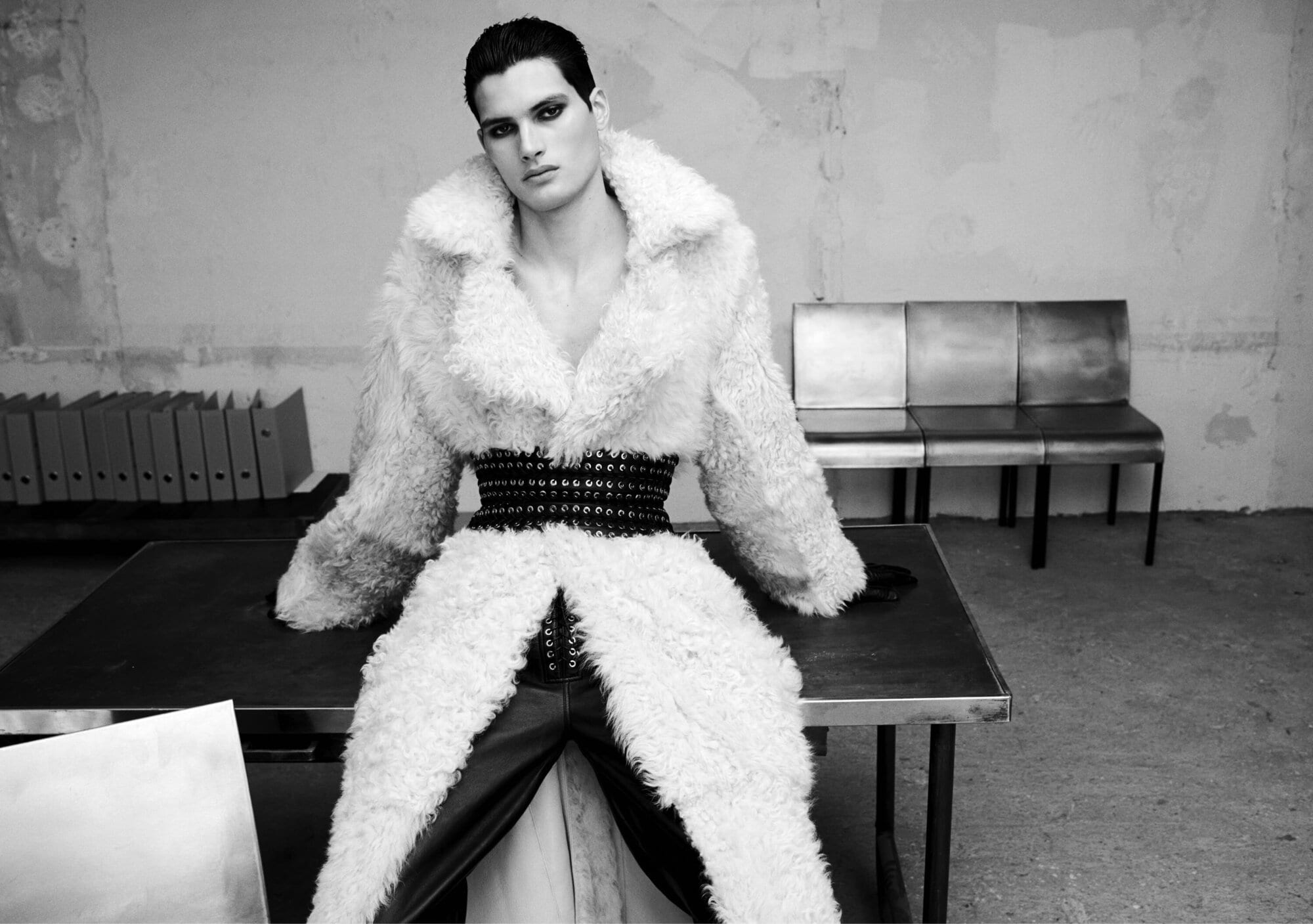 Model in a fluffy white textured coat and studded belt seated on a table in an interior space